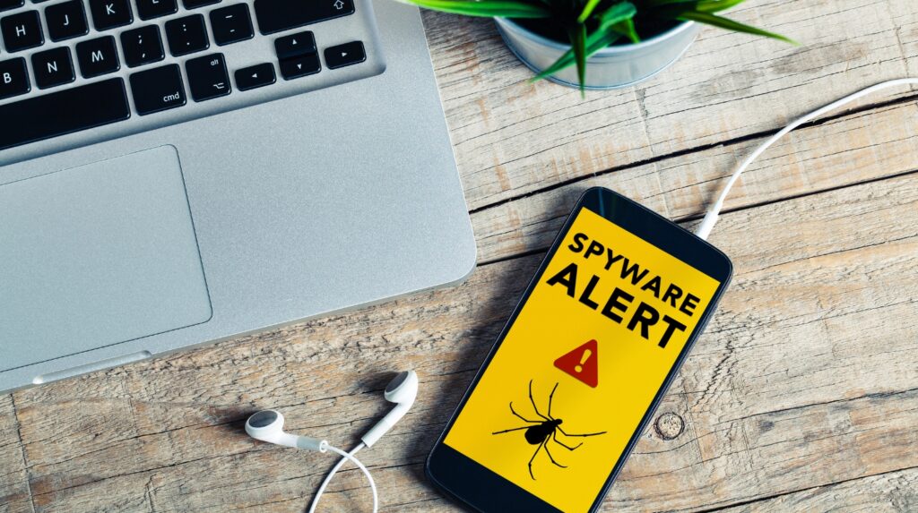 A smartphone on a wooden table displaying a yellow screen that reads “Spyware Alert” with a warning icon and bug graphic, placed next to a laptop, white earbuds, and a small plant.
